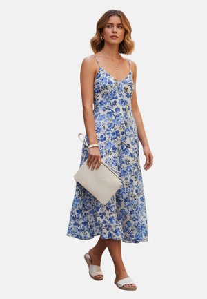Donna con abito midi senza maniche blu e bianco a fiori che tiene una pochette beige, indossa sandali bianchi a ciabatta e gioielli d'oro, in piedi su sfondo bianco.