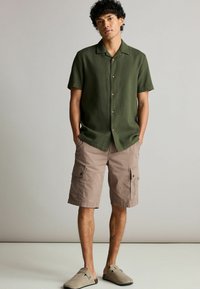 Mørkegrøn kortærmet tekstureret skjorte med træknapper, parret med lysebeige cargo shorts med lommer og grå slidesko.