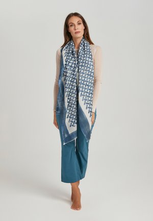 Sciarpa blu con motivo a disegni intrecciati, bordi con frange, drappeggiata elegantemente. Modello con indosso una maglietta chiara e pantaloni verde smeraldo, a piedi nudi, su uno sfondo neutro.