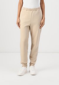 Beige joggingbroek met een geplooid elastische tailleband, taps toelopende benen en een zachte textuur. Gecombineerd met witte sneakers.