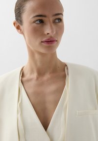 Blazer de color crema, texturizado, con un escote en V profundo y detalles de borde sin terminar. Presenta un pequeño bolsillo en el pecho y una tela ligera.