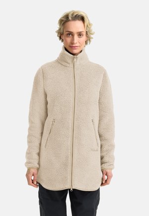 Donna che indossa una giacca sherpa in pile beige con cerniera, collo alto e tasche laterali con cerniera, in piedi davanti a uno sfondo bianco semplice.