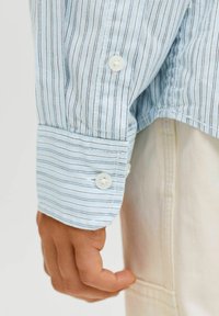 Světle modrá košile s dlouhým rukávem a tenkými vertikálními pruhy, se zapínáním na bílé knoflíky na manžetách, vyrobena z bavlny a s měkkou texturou.