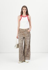 Débardeur blanc avec des accents rouges, associé à un pantalon large à imprimé léopard. Le modèle tient un petit sac à main gris clair. Fond blanc simple.