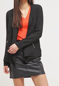 Veste noire cintrée avec ourlet asymétrique et poches zippées, portée sur un haut col en V orange vif, associée à une mini-jupe en cuir noire.