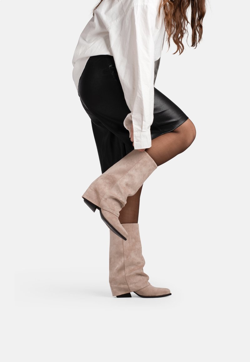 Woman wearing black skirt and white shirt adjusting beige suede mid-calf boots with pointed toes and low heels.