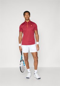 Red short-sleeve polo shirt with yellow accents, vertical stripes, and three buttons. Worn with white shorts, holding a tennis racket.