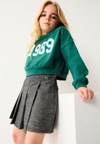 Zelený krátky sveter s potlačou "1989", kombinovaný s šedou sukňou s plisovaním v houndstooth vzore. Detaily zahŕňajú manžety a gombíkové akcenty.