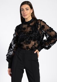 Zwarte doorschijnende blouse met bloemengalerij, hoge kraag en lange mouwen, gecombineerd met strakke zwarte broeken. Kenmerkt zich door textuurstof en gedetailleerde accenten.