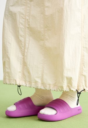 Purple rubber slides with a textured band design, worn over cream-colored socks, paired with a light beige skirt. Green floor.