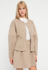 Bež jakna s teksturom, kratka, s okruglim izrezom, središnjim zatvaranjem na dugme i oversized rukavima, uparena s odgovarajućom suknjom.