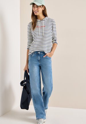 Kvinde iført lyseblå kasket, stribet langærmet hættetrøje, blå jeans, hvide sneakers, holder sort tote taske, står op ad beige væg.