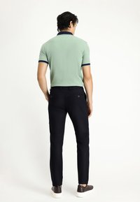 Lichtgroene polo met marineblauwe accenten, gecombineerd met zwarte broeken. Het model wordt van achteren getoond, met grijze sneakers.