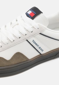 Hvid sneaker med en tekstureret brun ruskindsakcent, der har et blåt og rødt logo-patch, rund tå og hvide snørebånd.