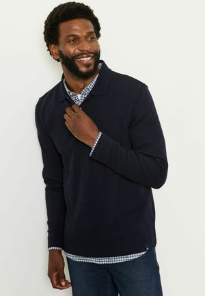 Lächelnder Mann, der ein marineblaues langärmeliges Polo über einem blau-weiß karierten Hemd und dunkelblauen Jeans trägt, vor einem hellen Hintergrund stehend.