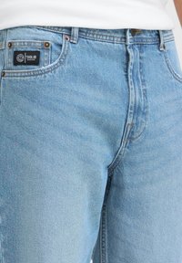 Vaqueros de mezclilla azul claro con un diseño clásico de cinco bolsillos, costuras doradas y una etiqueta negra cosida en el bolsillo trasero.