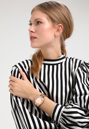 Femme aux cheveux blonds en queue de cheval, portant un haut rayé noir et blanc et une montre en or rose, regardant vers la gauche.