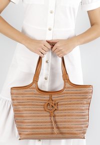 Tan geweven tote bag met een gladleren rand, voorzien van een gegraveerd logo en een kwastdetail, vastgehouden door twee stevige handvatten.