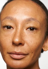 Nærbilde av et ansikt med glatt hud, fregner, mørkebrune øyne og naturlig leppefarge; håret trukket tilbake, nøytral bakgrunn.