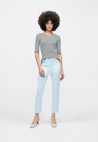 Haut à rayures noires et blanches à manches trois-quarts, associé à un pantalon bleu clair ajusté et des talons gris. Le mannequin est debout, les bras détendus.