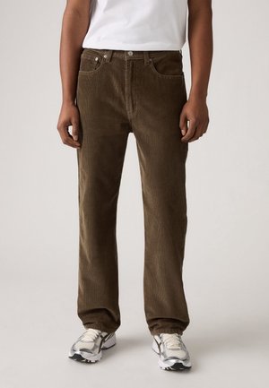 Homme portant un pantalon en velours côtelé marron, des baskets blanches et un t-shirt blanc, debout devant un fond clair uni.