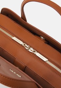 Ruskea nahkainen käsilaukun sisus, jossa vetoketjukiinnitys, kullanväriset metalliosat ja sileä pinta, sisältäen sisätaskun ja brändäyksen.