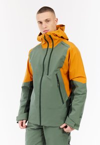 Waterdichte jas in groene en oranje kleurvlakken. Beschikt over een hoge kraag, ritszakken en een verstelbare capuchon voor extra functionaliteit.