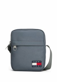 Grå rektangulær taske lavet af stof, med lynlås, justerbar strop og et logo-patch med røde, hvide og blå elementer.