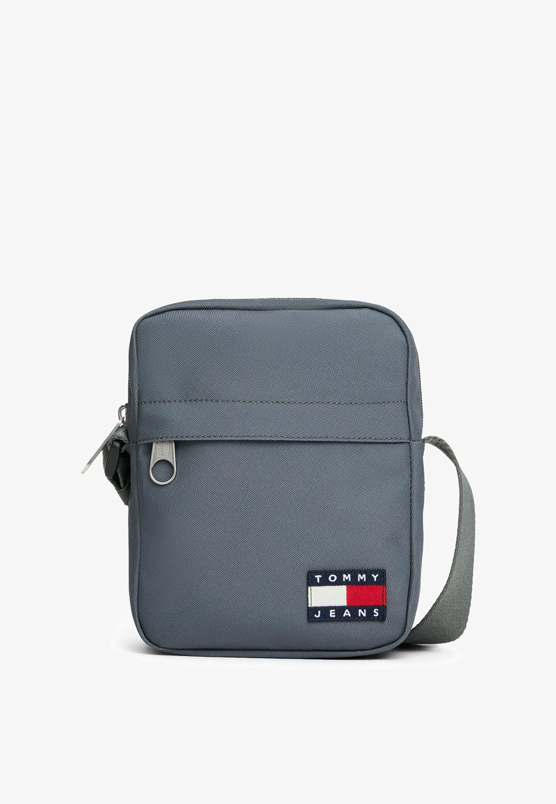 Graue rechteckige Tasche aus Stoff, ausgestattet mit einem Reißverschluss, einem verstellbaren Gurt und einem Logo-Patch mit roten, weißen und blauen Elementen.