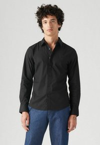 Camisa negra de botones ajustada, hecha de una tela suave. Presenta un cuello puntiagudo y mangas largas. Combinada con jeans azules.