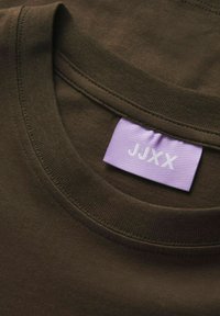 Hnědý bavlněný tričko s žebrovaným límcem. Na fialkovém štítku je bílými písmeny napsáno "JJXX". Hladká textura, žádné viditelné vzory nebo akcenty.
