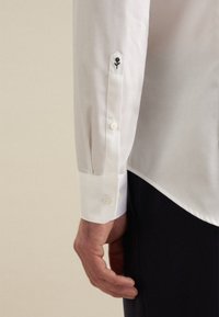 Witte shirt met een puntige manchet, voorzien van knoopsluiting en zwarte bloemenborduursels op de mouw. Glad textuur en een aangesloten ontwerp.