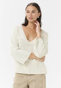 Pull en maille lâche beige clair avec un col en V profond, des manches larges en forme de cloche et un ourlet côtelé, assorti à un pantalon beige.