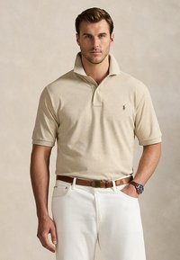 Mand iført beige poloshirt med opstående krave, hvide bukser, brun bælte og armbåndsur, stående mod ensfarvet baggrund.