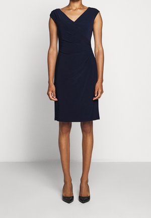 Femme portant une robe noire ajustée, longueur genou, manches courtes avec un décolleté en V et des talons noirs à bout pointu, debout devant un fond uni.