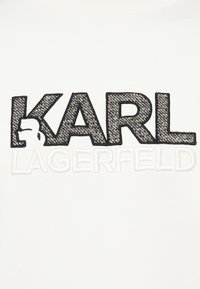 Vyšívaný text "KARL" v čiernej a štruktúrovanej hnedej farbe s malou bielou siluetou na písmenku K, nad bielo vyšívaným "LAGERFELD" na bielom látkovom pozadí.