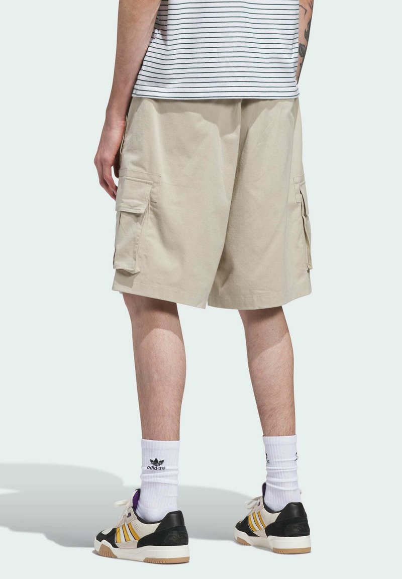 Persona que lleva pantalones cortos cargo beige, calcetines blancos Adidas, zapatillas negras y amarillas, y una camisa a rayas, vista desde atrás.