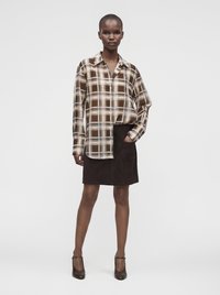Femme portant une chemise à carreaux marron et beige boutonnée, une jupe marron foncé avec poche et des talons marron foncé, debout avec une main dans la poche.