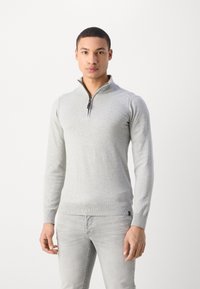 Pull en maille gris clair avec un col zippé moitié, des poignets et un ourlet côtelés, présentant une texture lisse et un design ajusté.