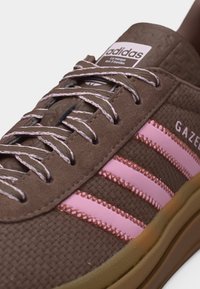 Gros plan sur une basket Adidas Gazelle marron avec des rayures roses, des lacets à motifs, une tige texturée et une semelle gomme.