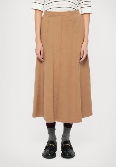 Femme portant une jupe midi couleur camel, des chaussettes grises avec des bordures bordeaux, des mocassins noirs avec un détail en chaîne dorée, et un haut blanc à motifs.