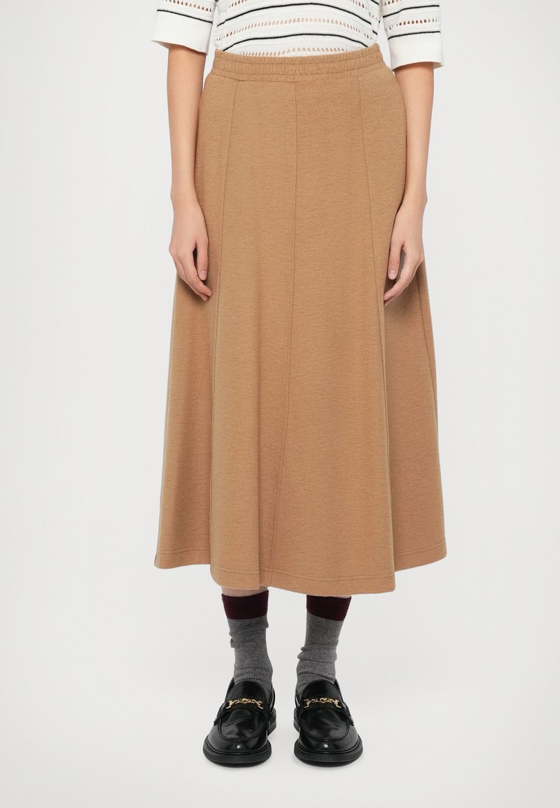 Donna che indossa una gonna midi color cammello, calze grigie con parte superiore bordeaux, mocassini neri con dettaglio in catena dorata e una maglietta bianca a fantasia.