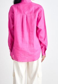 Långärmad, rosa linneskjorta med en bakveck och knäppte ärmslut, parat med krämfärgade cargobyxor, som visas bakifrån.