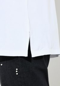 Weißes T-Shirt mit seitlichen Schlitzen, aus weichem Stoff gefertigt. Es wird über dunklen Denim-Hosen mit Perlenakzenten an den Taschen getragen.