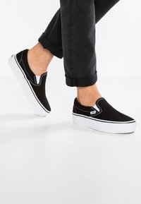Zapatillas de lona negra sin cordones con suelas blancas, costuras blancas en contraste y un diseño de perfil bajo. Presenta un clásico logo de Vans.