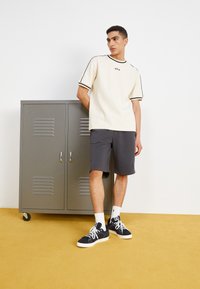 Creme kortærmet skjorte med sort kant, grå shorts og sorte sko med hvide såler. En grå opbevaringsboks i baggrunden.