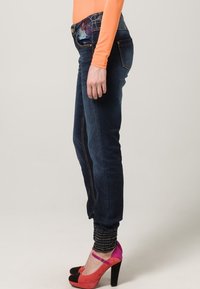 Henkilö, jolla on yllään tummansiniset farkut, joissa on kukkakirjontaa taskussa, oranssi pitkähihainen paita sekä vaaleanpunaiset ja mustat korkokengät.