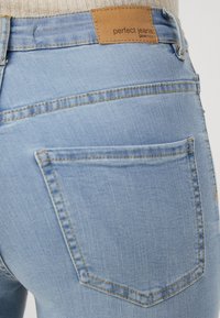 Jeans de ganga azul claro com um design ajustado, apresentando uma etiqueta de couro bege na cintura e bolsos traseiros standard.