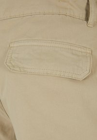 Los pantalones de algodón beige presentan un bolsillo trasero rectangular con un contorno cosido, una textura suave y un sutil patrón de trama diagonal.