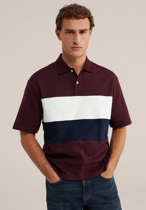 Homme aux cheveux bouclés portant un polo à manches courtes avec des rayures horizontales bordeaux, blanches et bleu marine, mains dans les poches du jean, fond neutre.